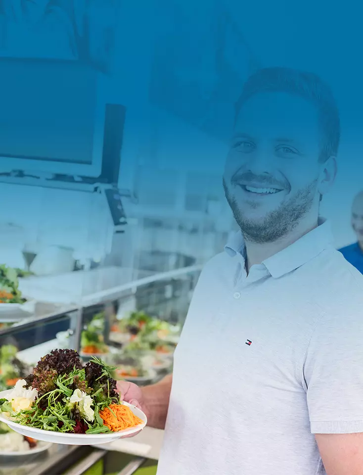 A man holding a plate of salad in a canteen.