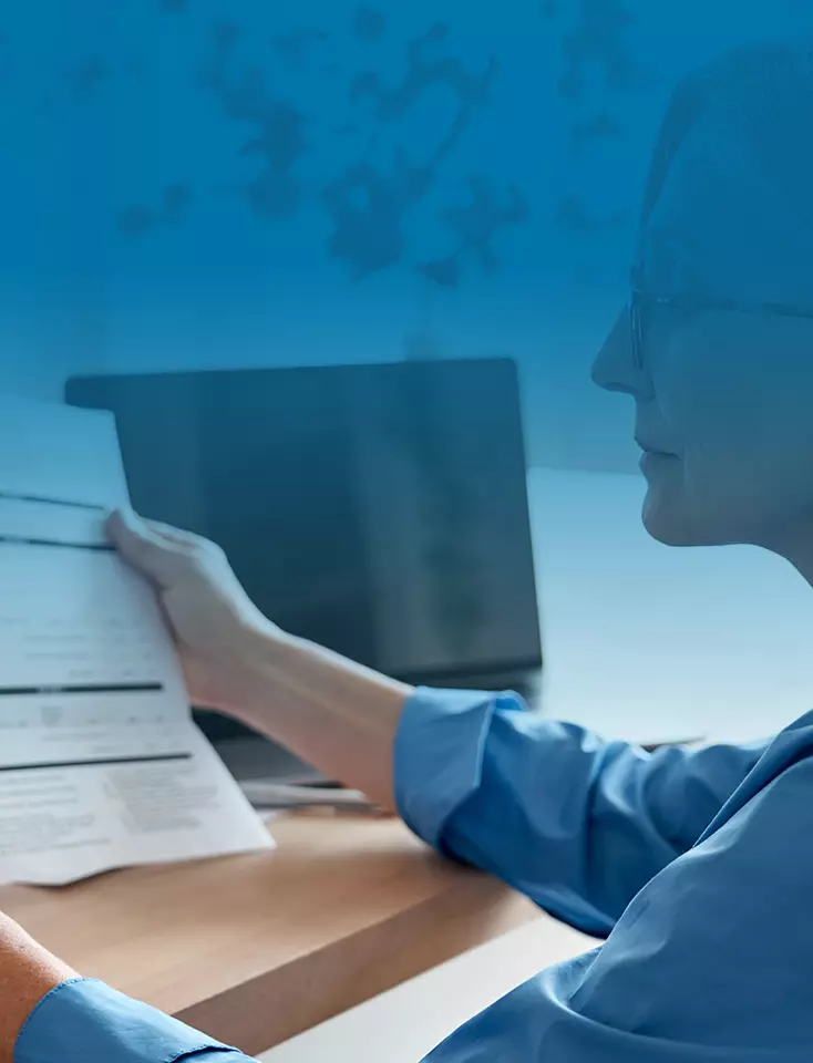 A woman sits in front of a laptop and holds a sheet of paper in her hand.